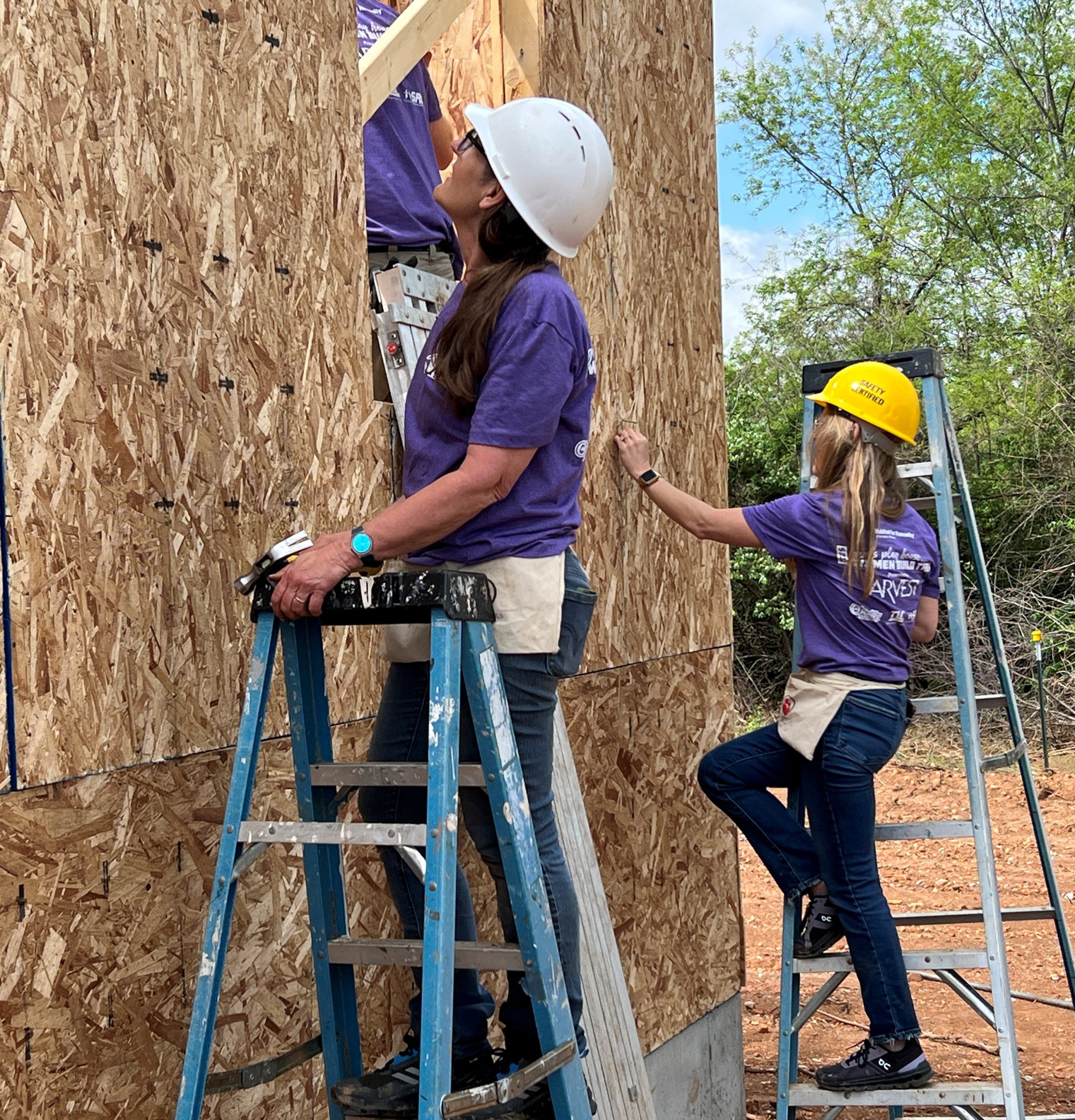 Two women in hard hats hammer nails into the exterior wall of a house that's under construction.