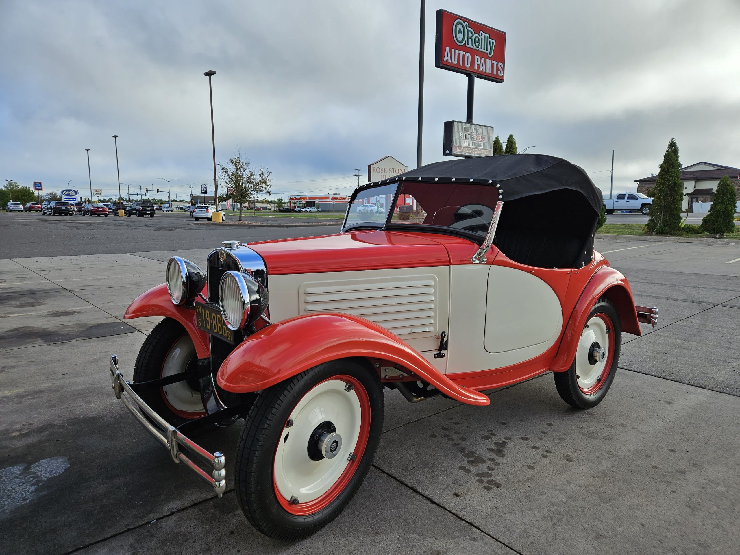 red and white 1931 American Austin Bantam roadster with black top, parked in front of O'Reilly Auto Parts sign