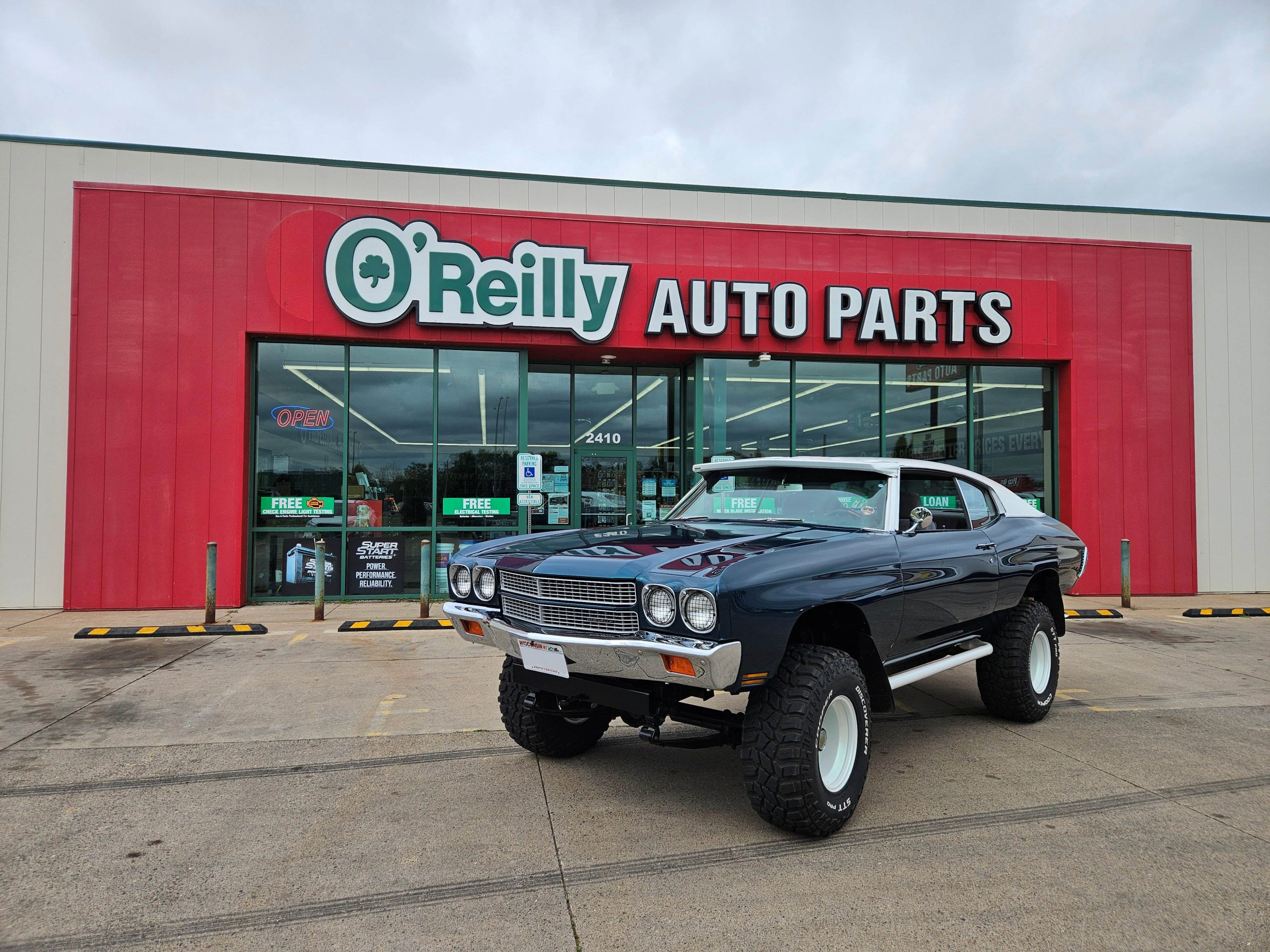 black vintage Chevelle on large tires parked in front of O'Reilly Auto Parts store on overcast day