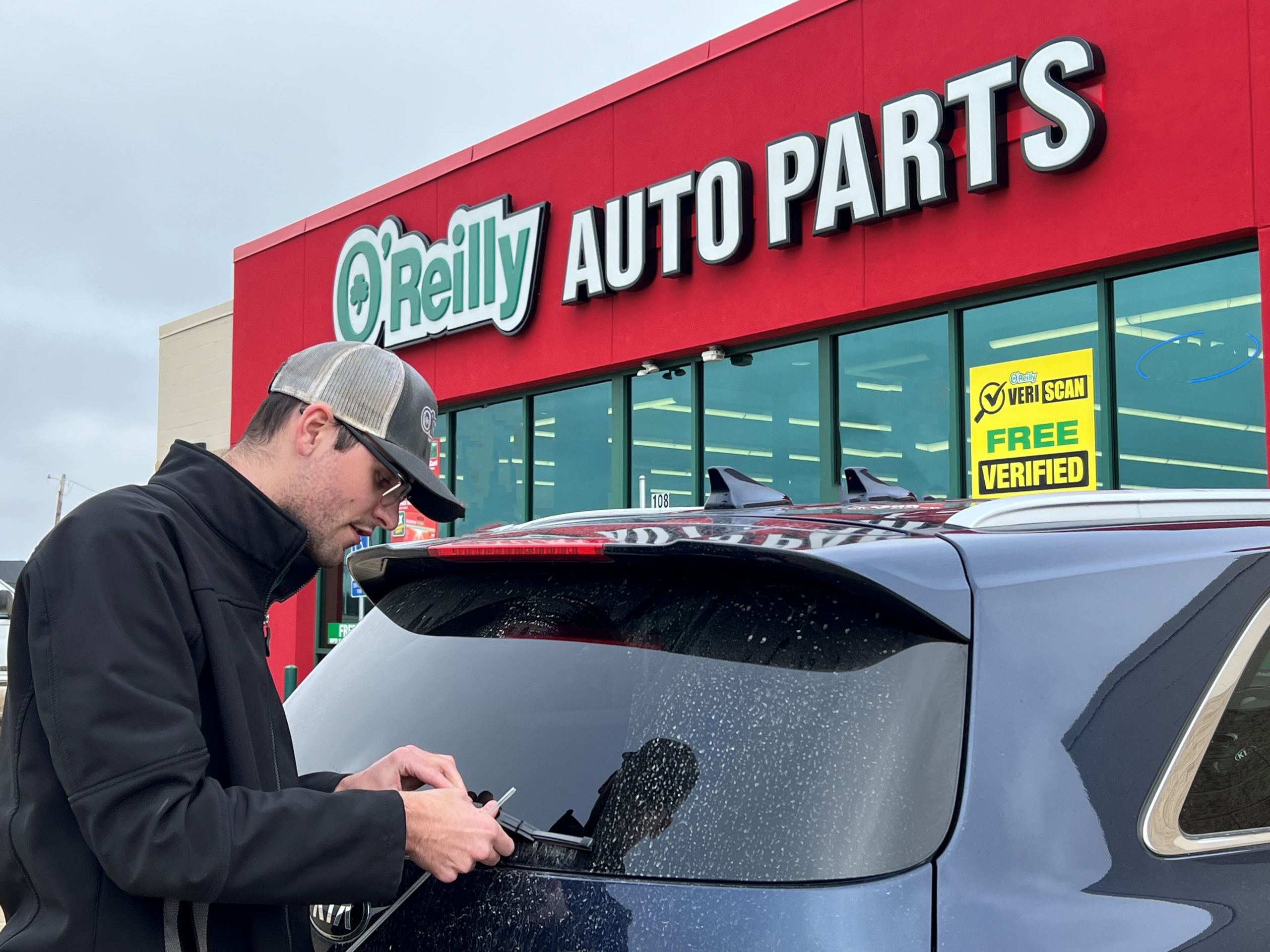 Image shows a man in a black coat installing a rear windshield wiper on a blue vehicle.